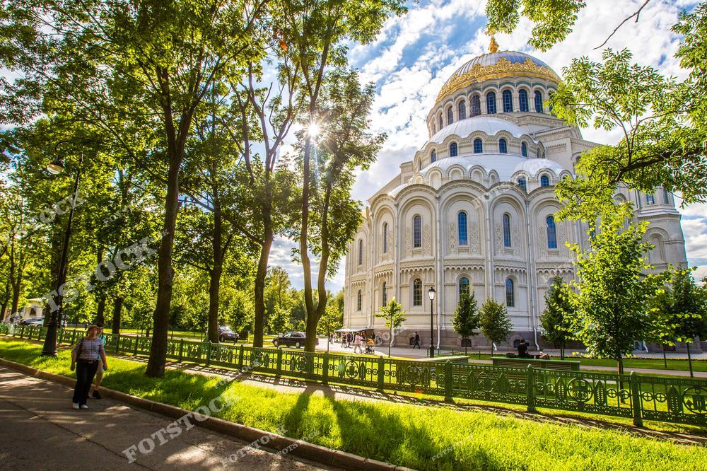 На фотографии показан Морской Никольский собор в окружении зелёных деревьев Кронштадта. Солнечный свет, проникающий сквозь листву, создает атмосферу тепла и уюта, подчёркивая архитектурные детали здания. Этот собор, возведённый в честь моряков, является важным духовным и культурным центром. Летний парк вокруг собора предлагает посетителям спокойное место для отдыха и прогулок, создавая гармоничное сочетание природы и религиозной архитектуры.